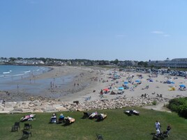 Uma praia com pessoas tomando sol e nadando, um estacionamento com carros e um edifício ao fundo.