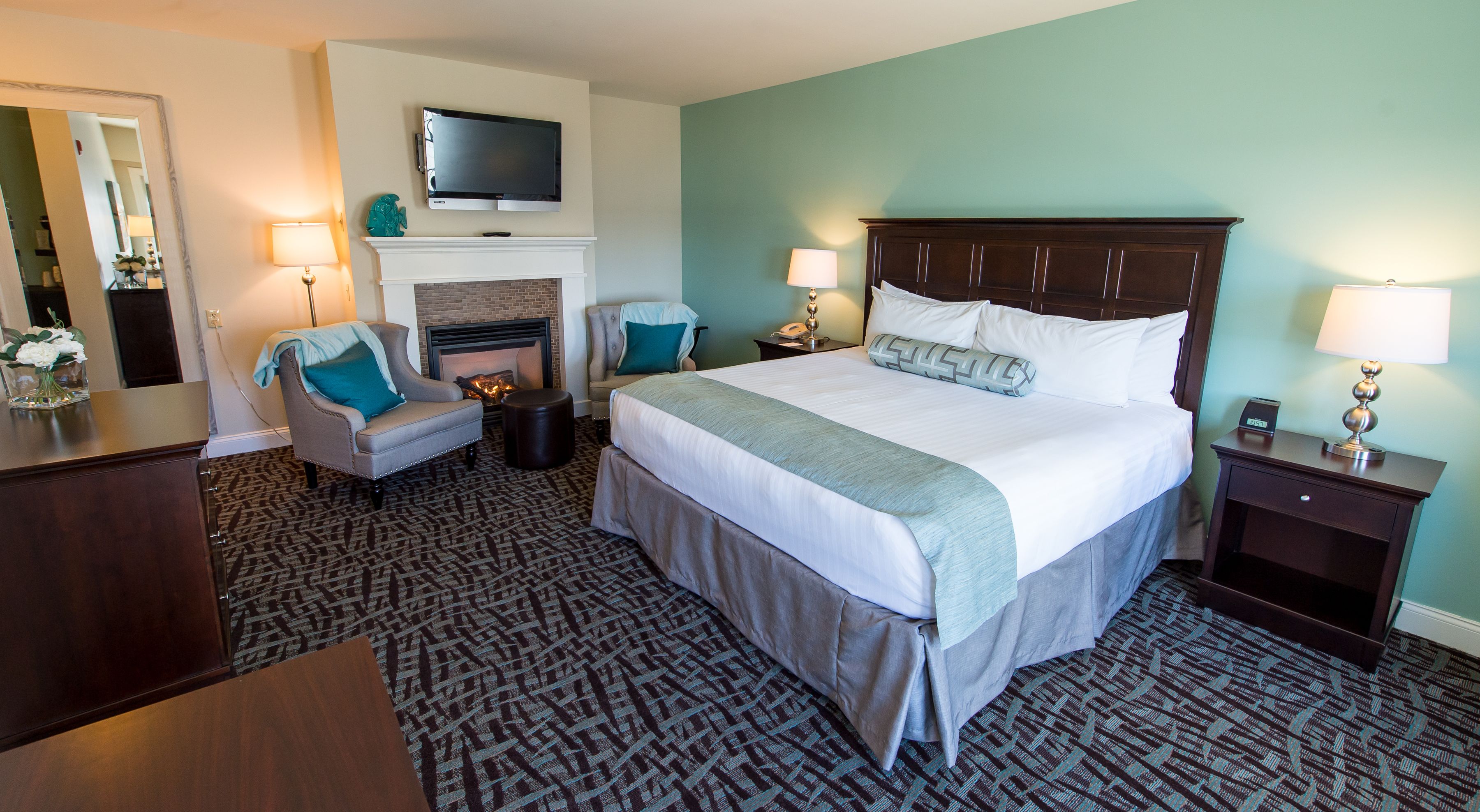 A hotel room with a large bed, two chairs, a fireplace, a TV mounted above the fireplace, and a nightstand with a lamp.