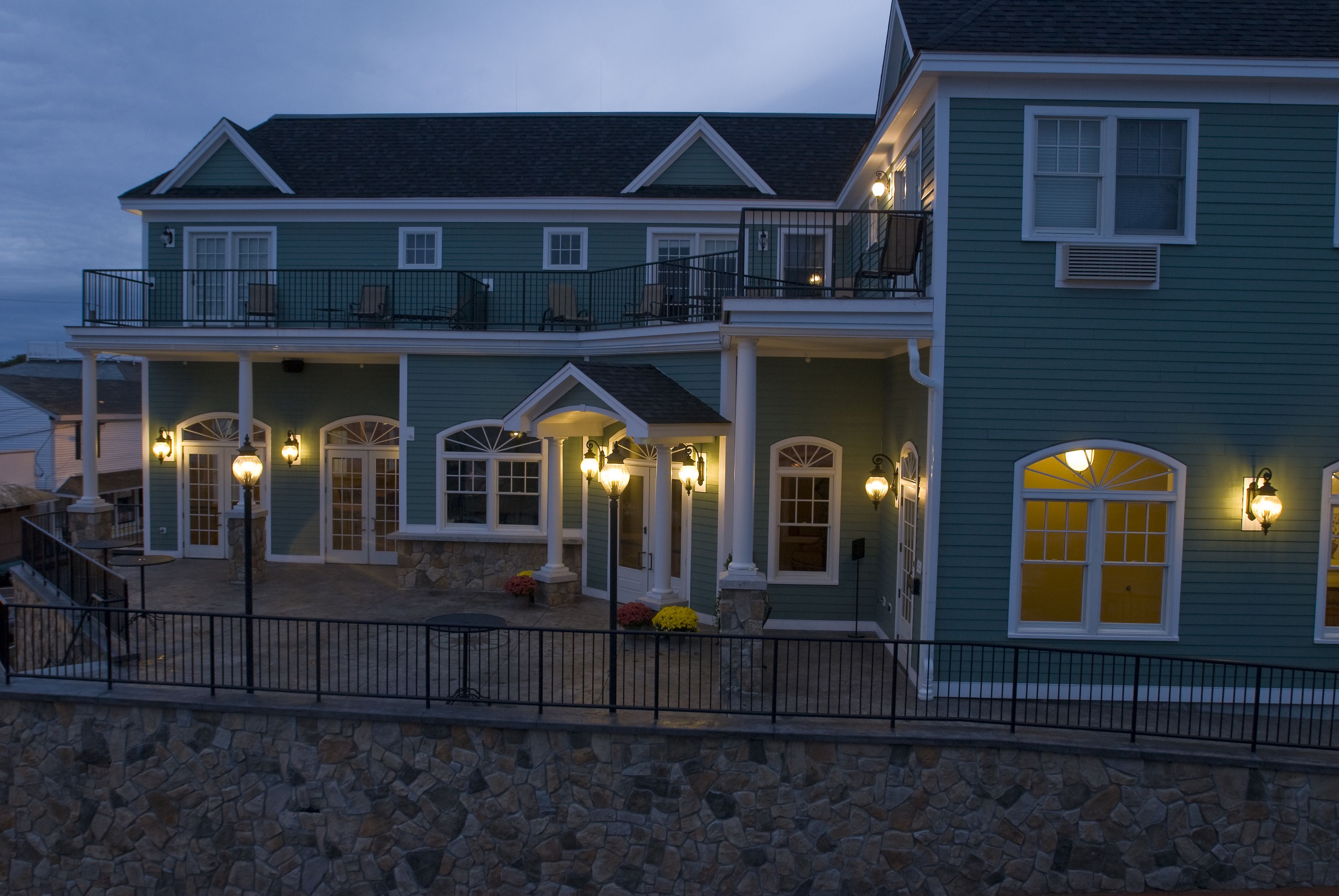 A two-story house with a balcony, stone foundation, and multiple windows with exterior lighting.