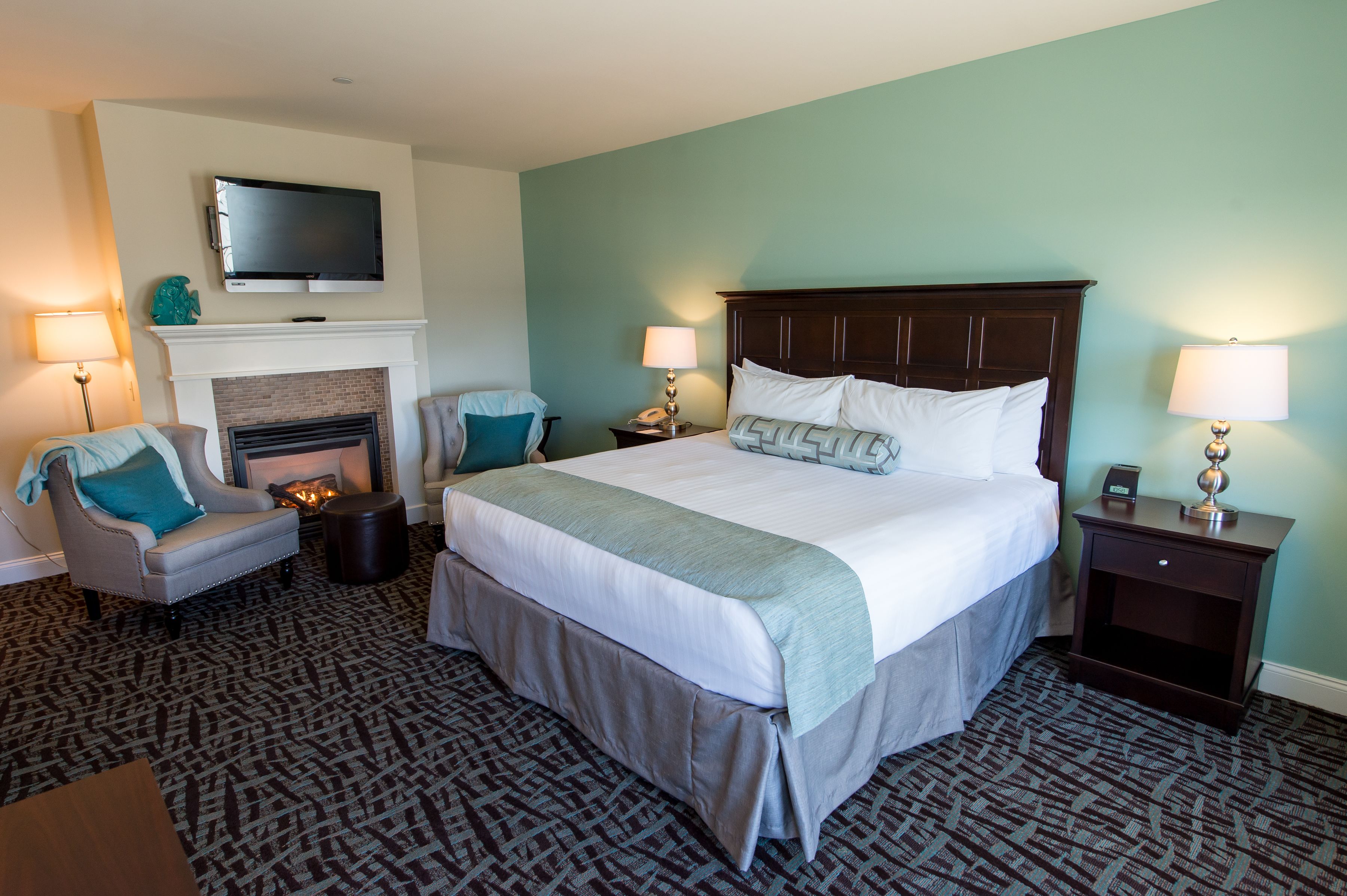 A hotel room with a large bed, a fireplace, a TV mounted on the wall, and two bedside tables with lamps.