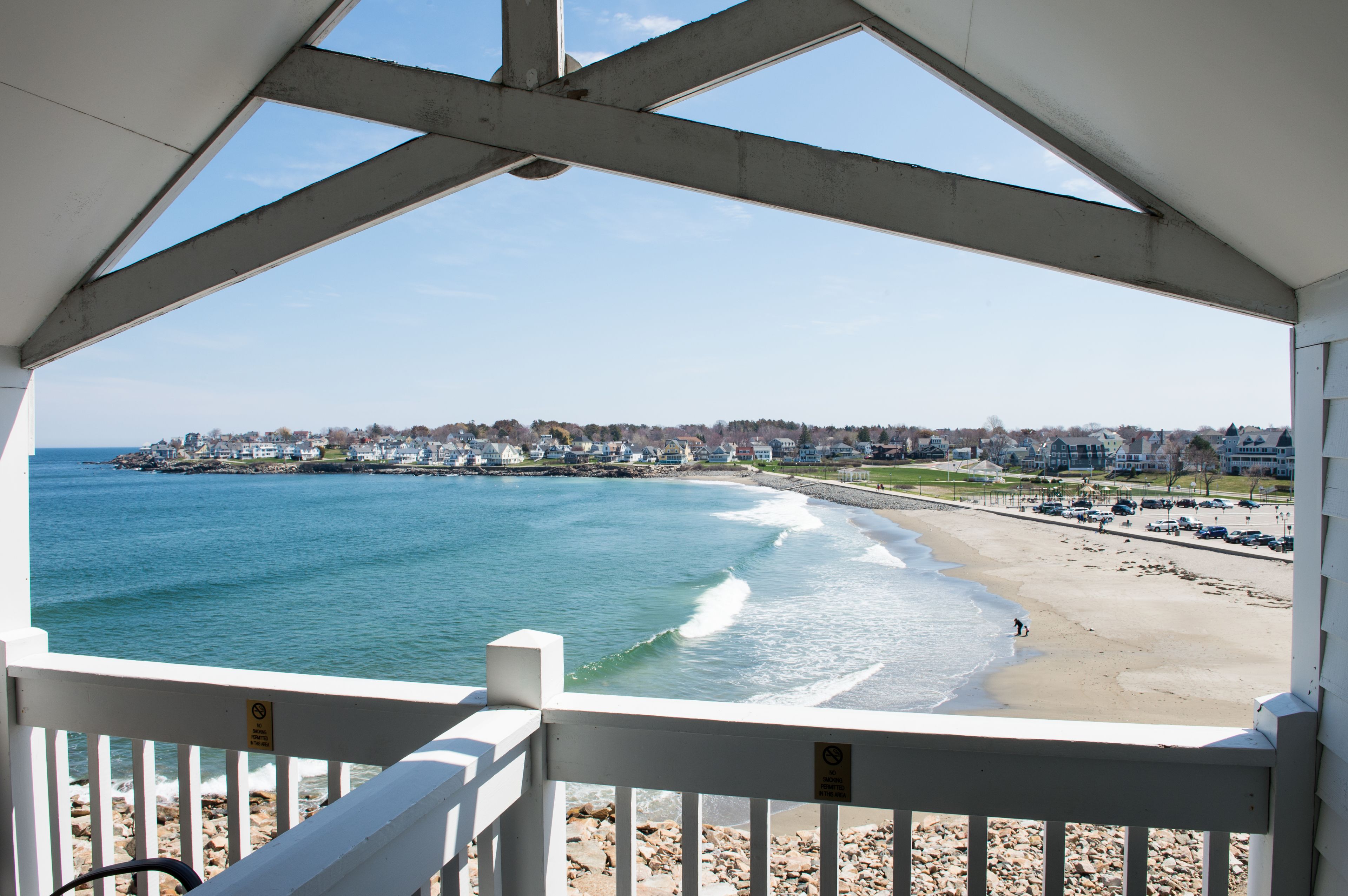 A beach view from a covered area with a railing, a clear sky, and a distant shoreline with buildings.