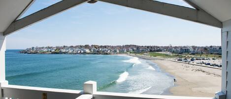 A beach view from a covered area with a railing, a clear sky, and a distant shoreline with buildings.