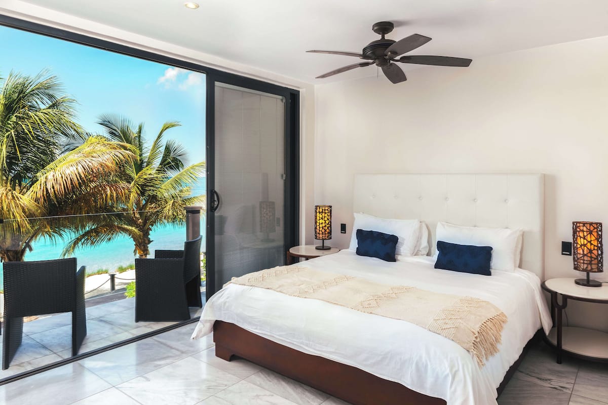 A bedroom with a large bed, two nightstands, a ceiling fan, and a view of palm trees and the ocean through a sliding glass door.