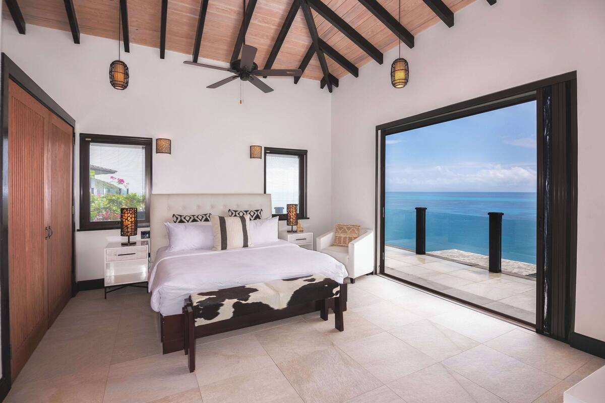 A bedroom with a bed, bedside tables, a nightstand, a ceiling fan, and a large window with a view of the ocean.