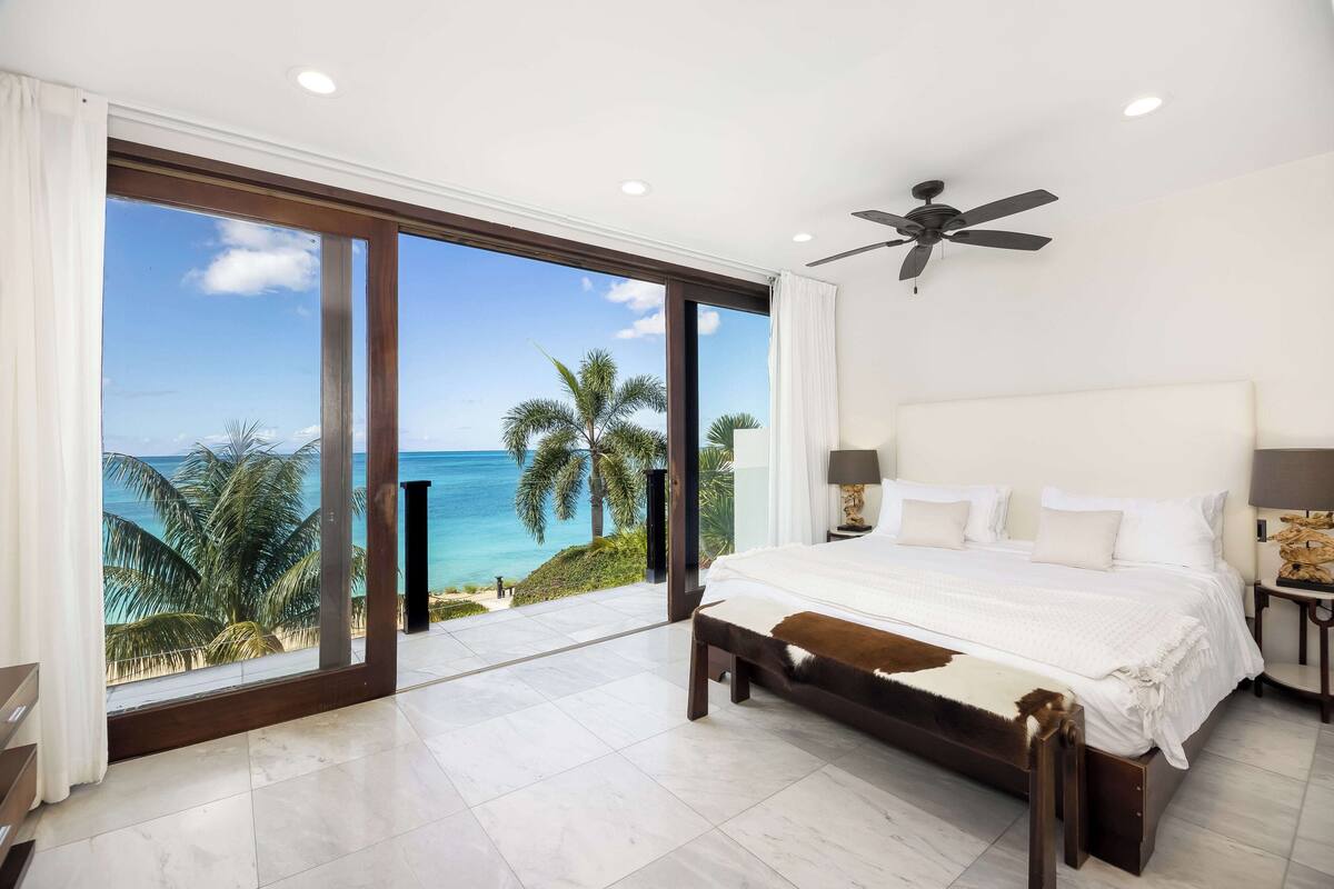 A bedroom with a large bed, a ceiling fan, and a view of the ocean through a sliding glass door.