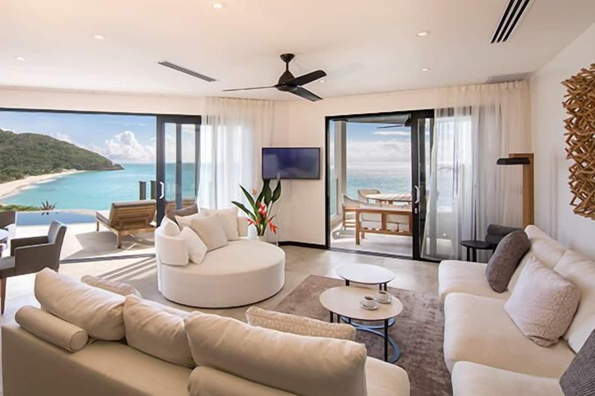 A modern living room with a large sofa, a round coffee table, and a dining area with a table and chairs. There is a television mounted on the wall and a ceiling fan. The room has large windows with a view of the beach and ocean.