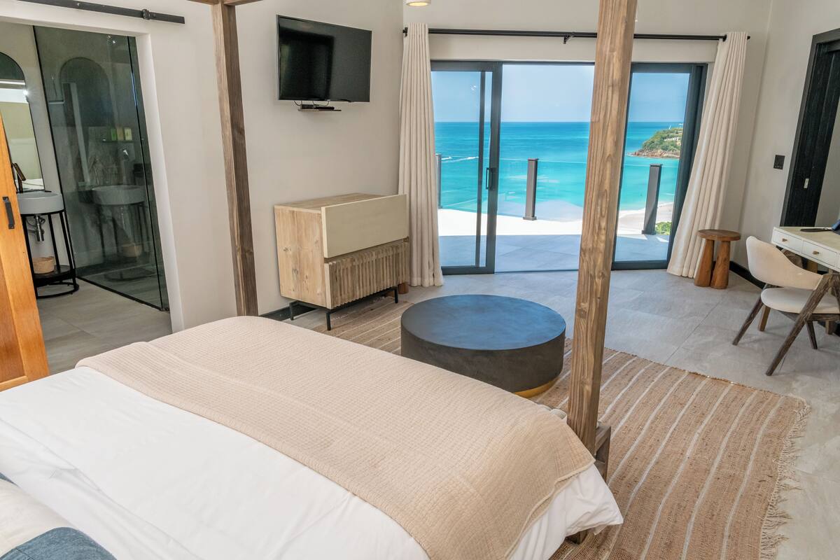 A bedroom with a large bed, a wooden nightstand, a television, and a view of the ocean through a sliding glass door.