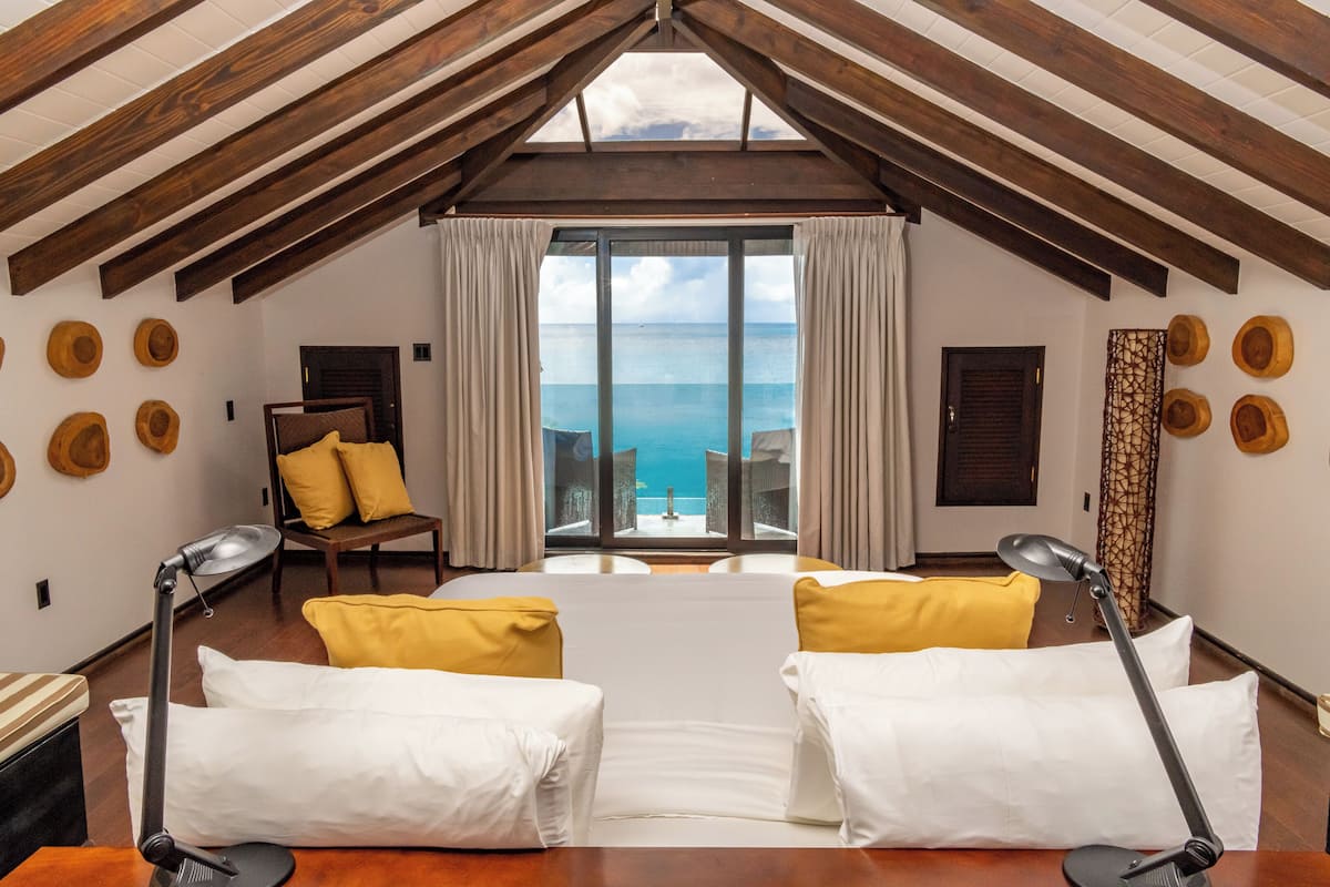 A room with a sloped ceiling, wooden beams, a large window with a view of the ocean, a bed with white linens, two yellow pillows, and a desk with a lamp.