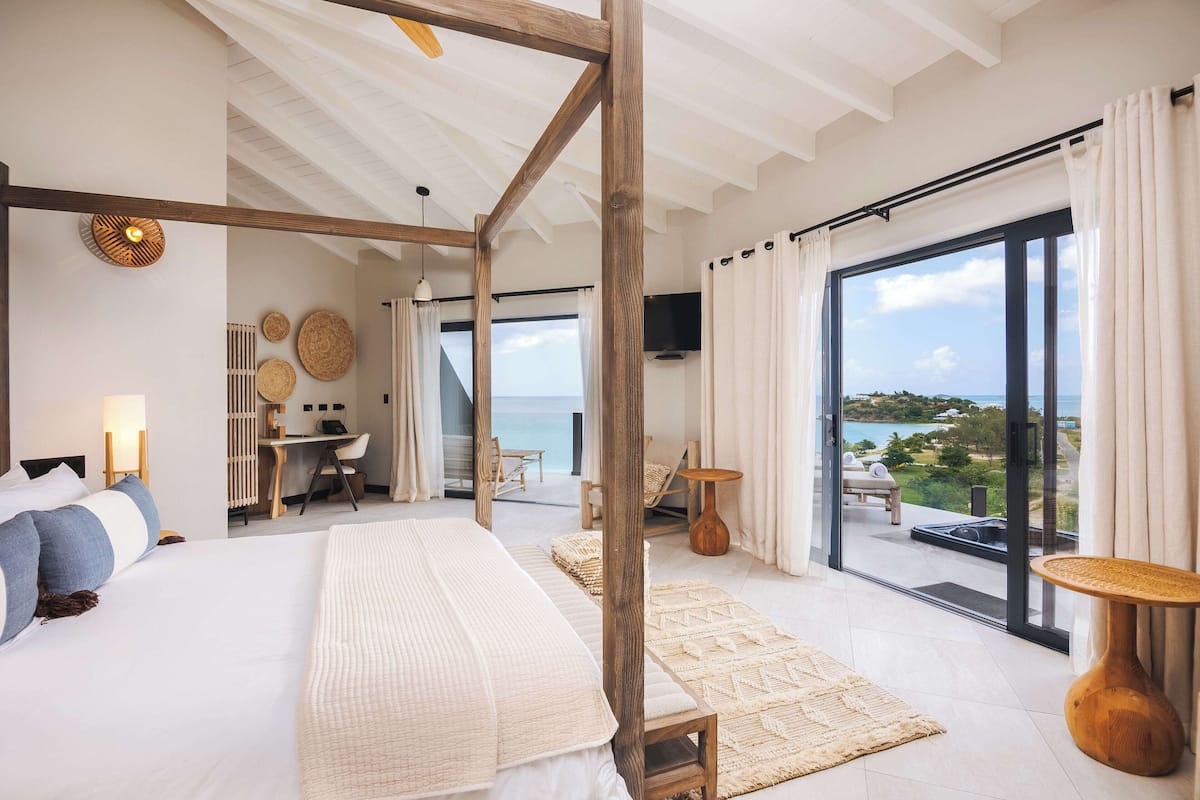 A modern bedroom with a large bed, a wooden ceiling, a desk, and a view of the sea through large windows.