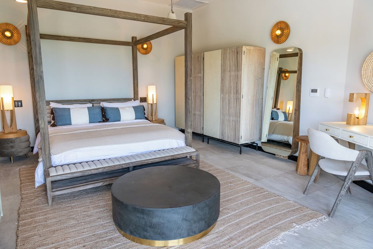 A spacious bedroom with a four-poster bed, a round ottoman, a wooden desk, and a large mirror.