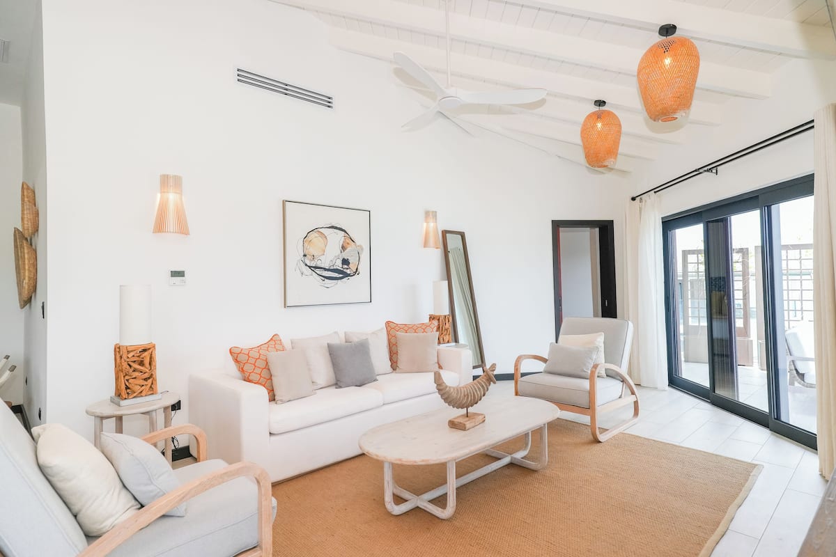 A modern living room with a white sofa, a coffee table, and a chair. There is a large window with curtains, a ceiling fan, and decorative wall art.