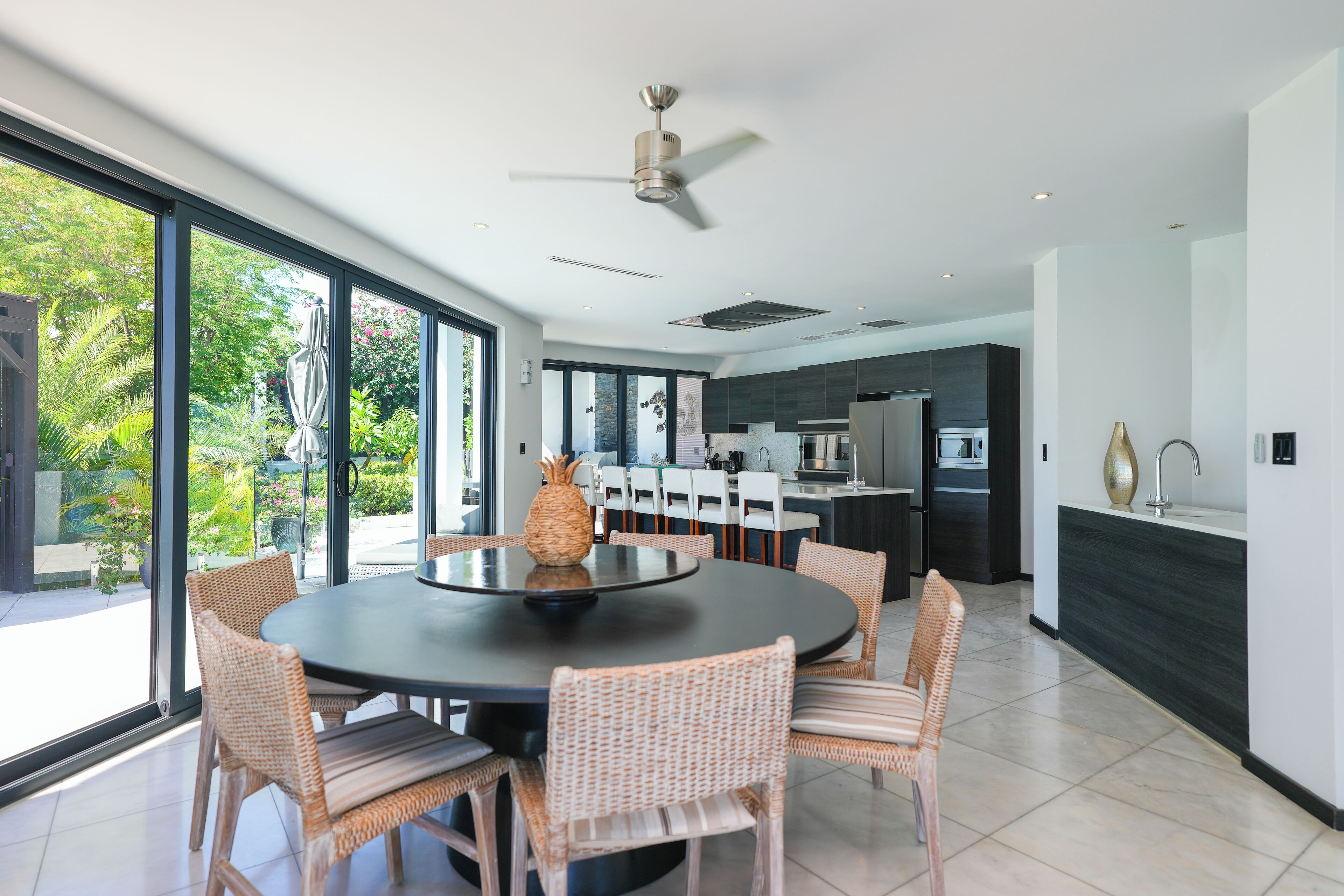 A modern kitchen with dark cabinetry, a central island, and a dining area with wicker chairs and a round table.