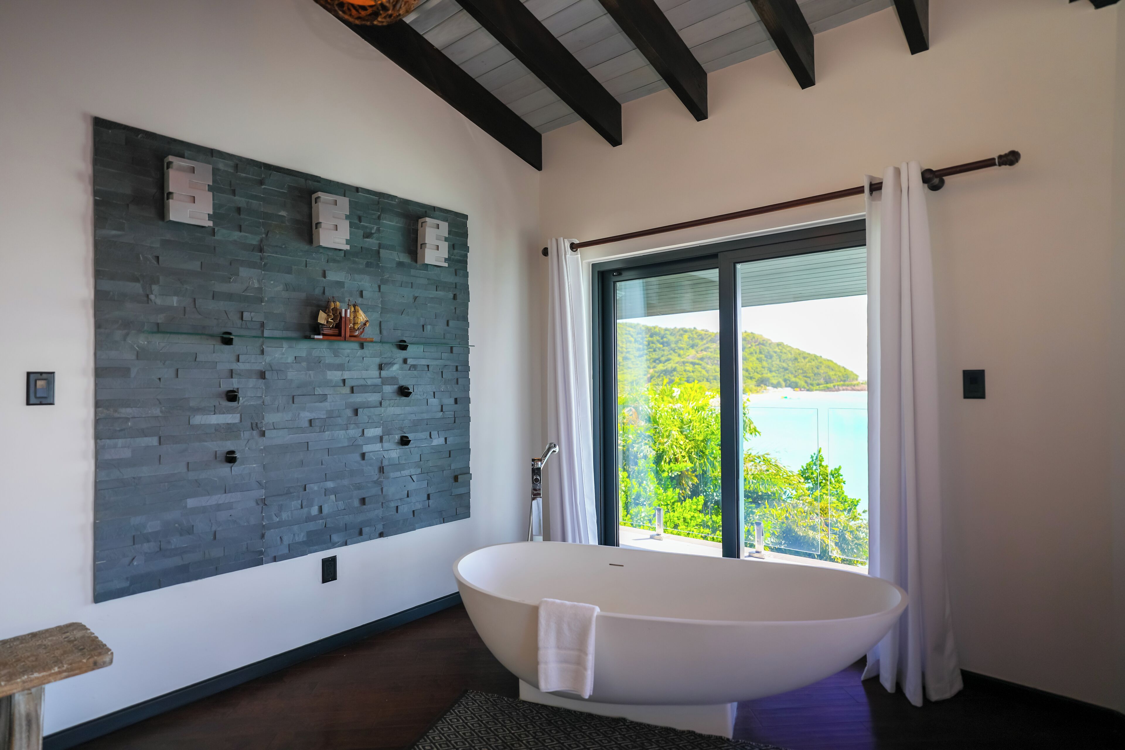 A modern bathroom with a freestanding bathtub, a stone wall, and a large window offering a view of a green landscape.