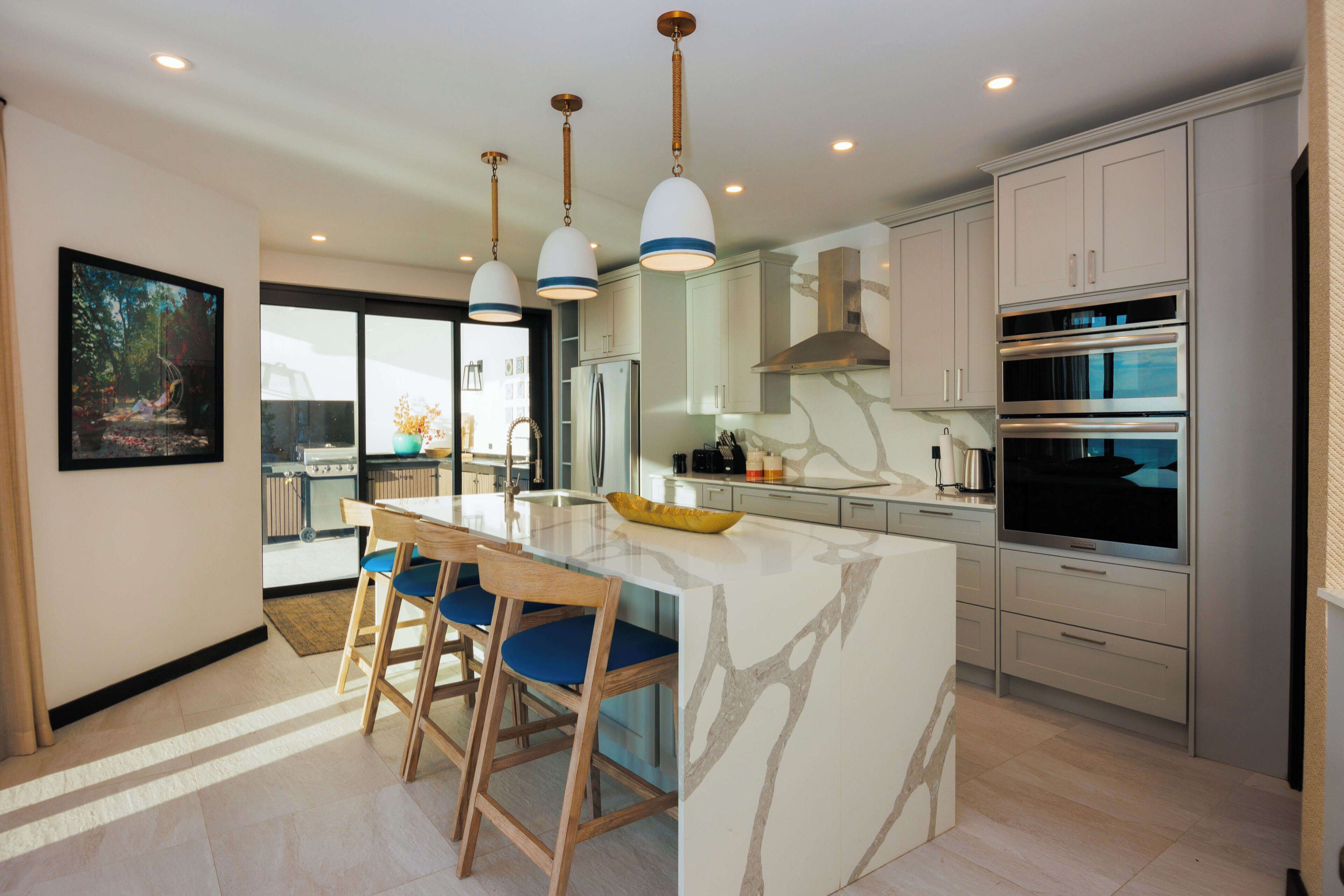 A modern kitchen with a central island, pendant lighting, stainless steel appliances, and a view to the outside.