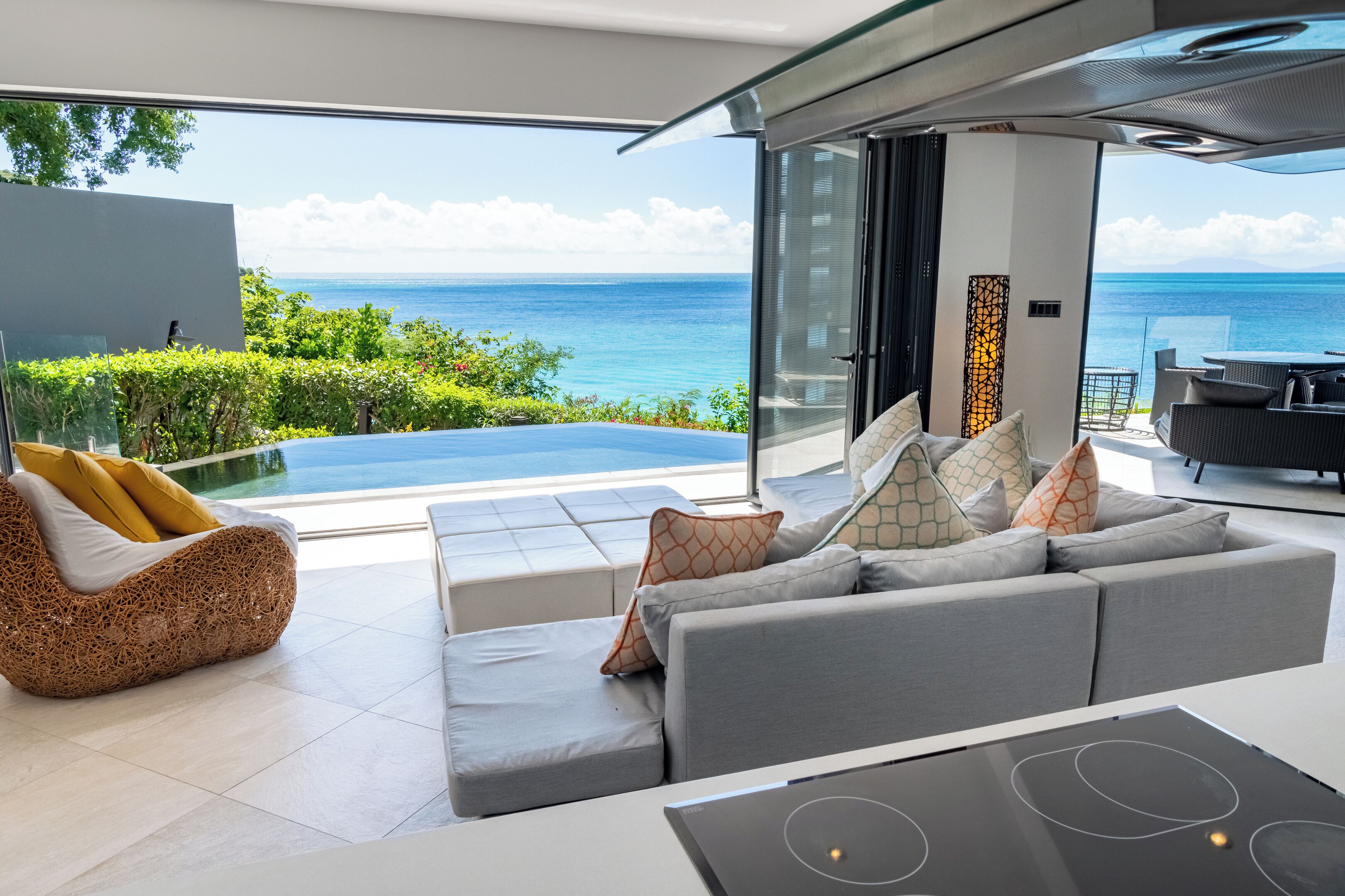 A modern living room with a grey sofa, a wicker chair, and a dining area. A large glass door leads to a balcony with a view of the ocean.