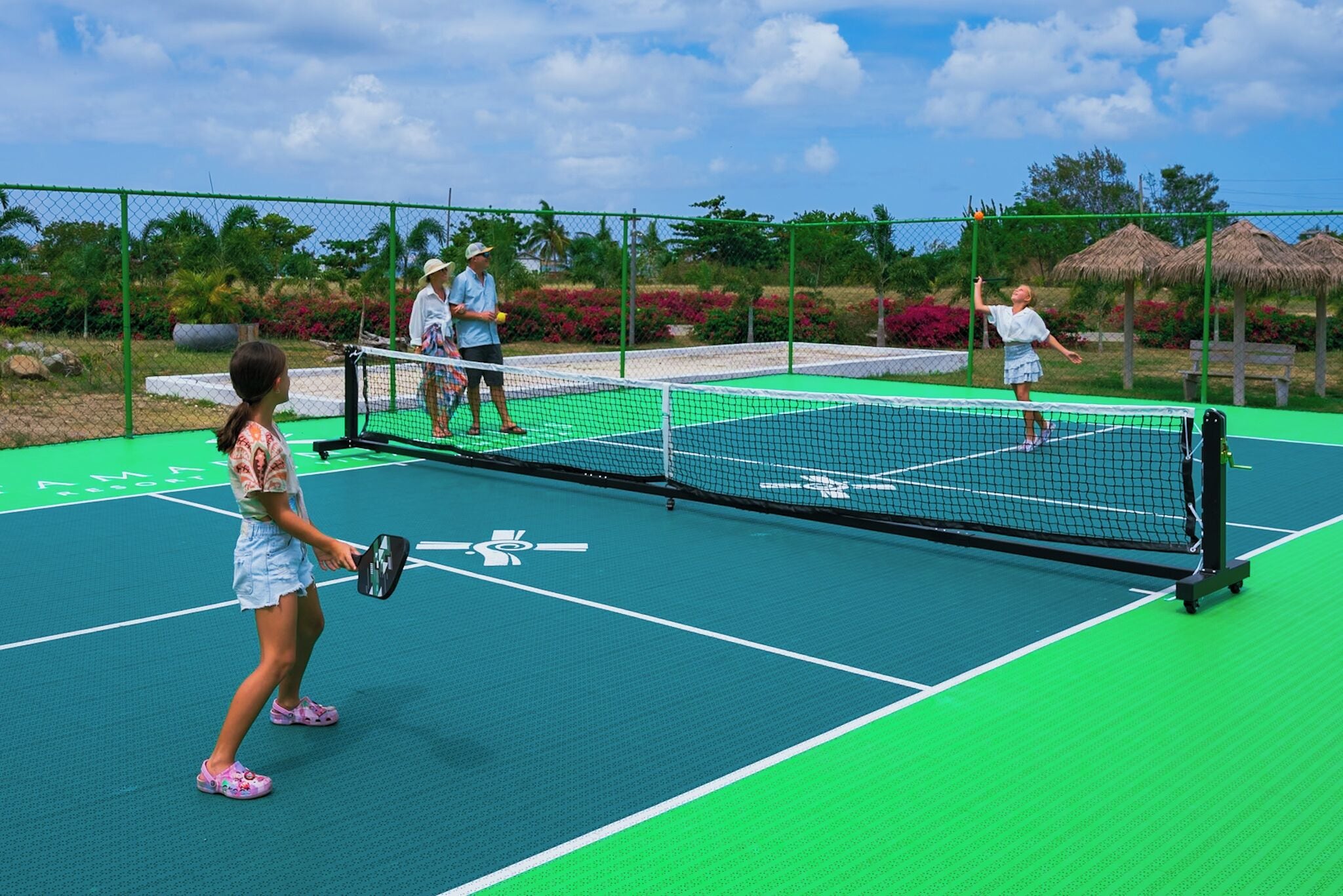 An outdoor tennis court with a net, surrounded by a green fence and tropical landscaping.