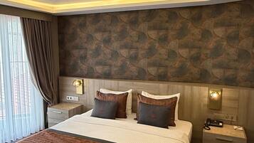 Une chambre d’hôtel moderne avec un grand lit, deux tables de chevet, une petite table, une lampe fixée au mur et une fenêtre avec des rideaux.