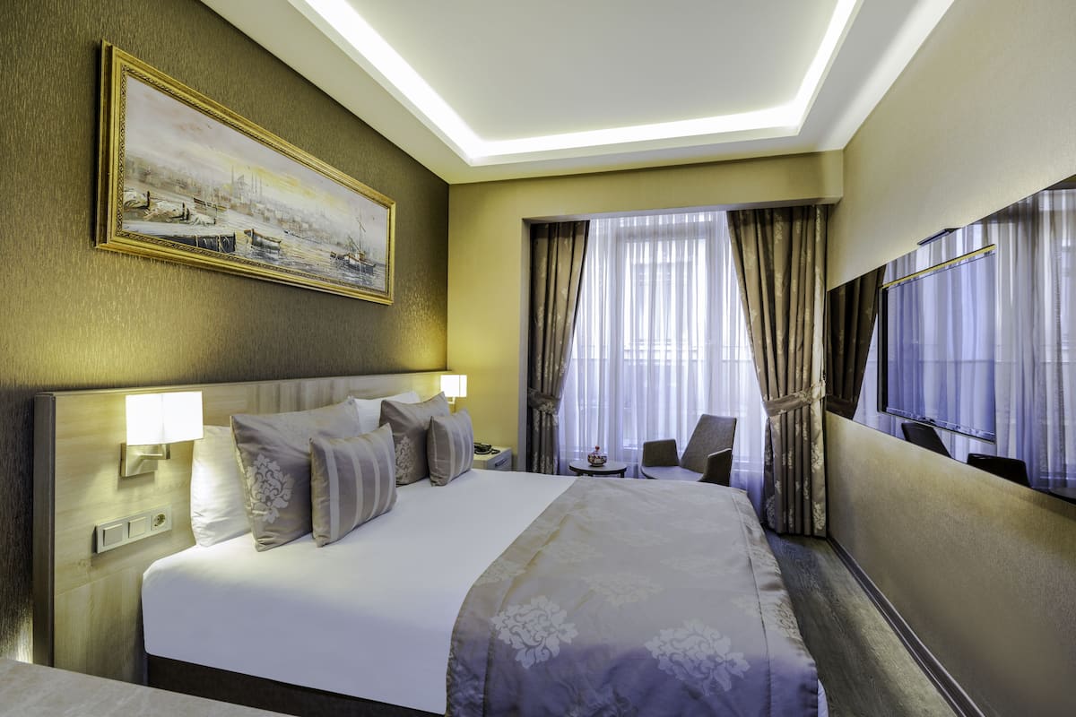 Habitación de hotel con una cama grande, un cuadro en la pared, un escritorio con una silla y un ventanal con cortinas.