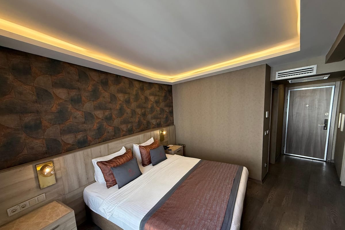 Una habitación de hotel moderna con una cama grande, cabecera con diseño de piedra, lámparas de noche y una mesita de noche de madera.
