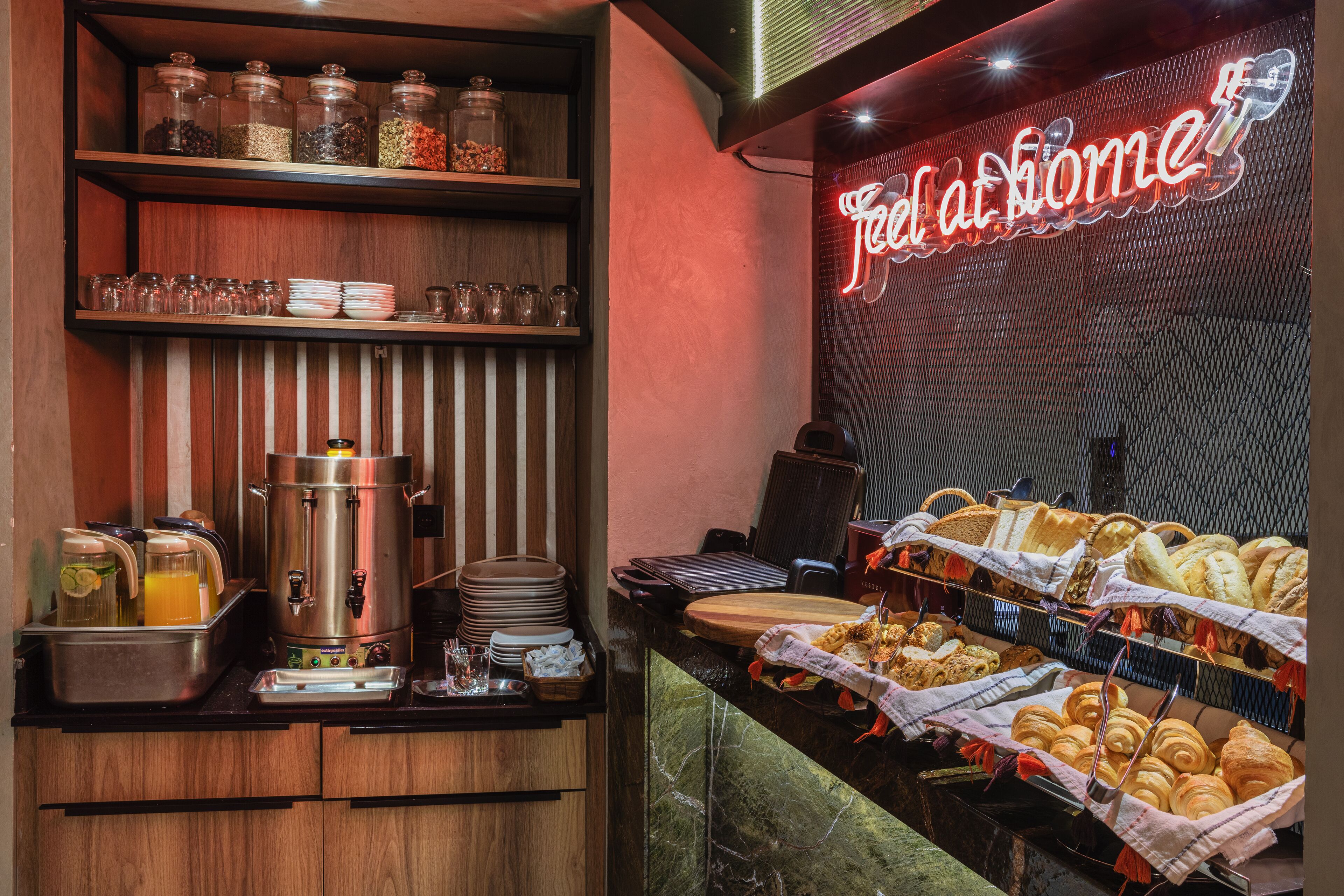 A breakfast buffet with various food items, a coffee machine, and a neon sign reading 'Feel at home'.