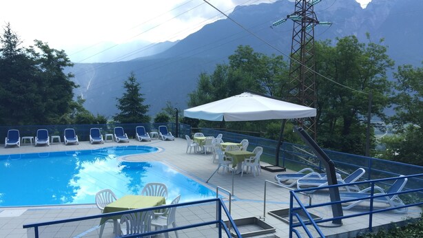 A swimming pool with lounge chairs and tables, surrounded by trees and mountains in the background. - Hotel Ristorante Scaranò (Levico Terme)