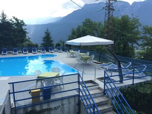 Une piscine entourée de chaises longues et de tables, avec des arbres tout autour et des montagnes en arrière-plan.