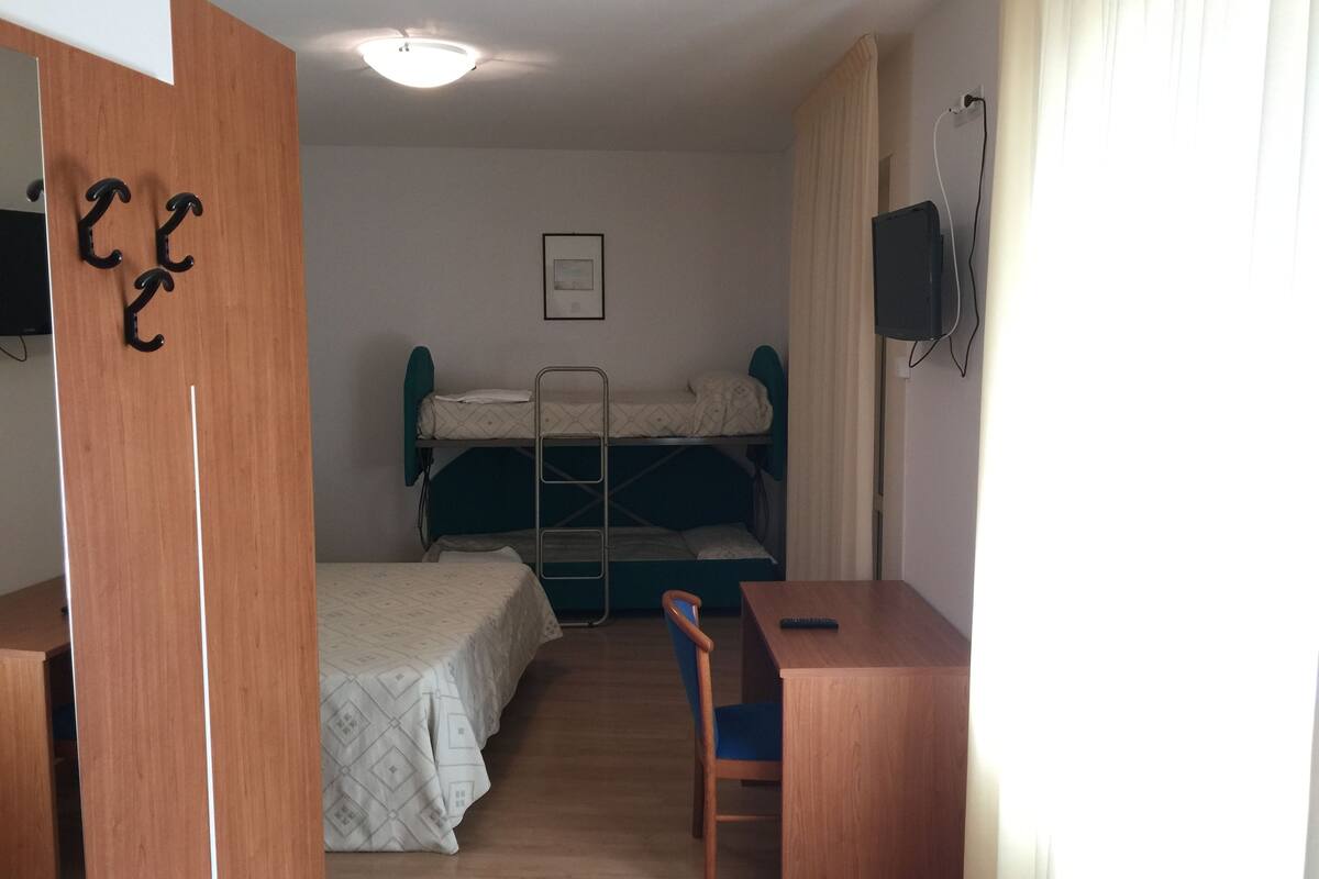 Ein kleines Zimmer mit Etagenbetten, einem Schreibtisch und einem Stuhl. Ein Fernseher ist an der Wand montiert und es gibt eine Deckenleuchte.