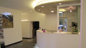 A reception area with a curved reception desk, a large mirror, a framed artwork on the wall, and a small flower arrangement on the desk. - Hotel Ristorante Scaranò (Levico Terme)