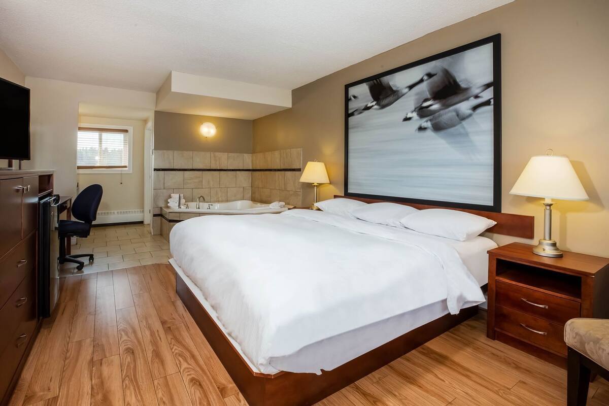 A hotel room with a large bed, a desk with a chair, a desk lamp, a wall-mounted TV, and a bathroom with a bathtub and sink.