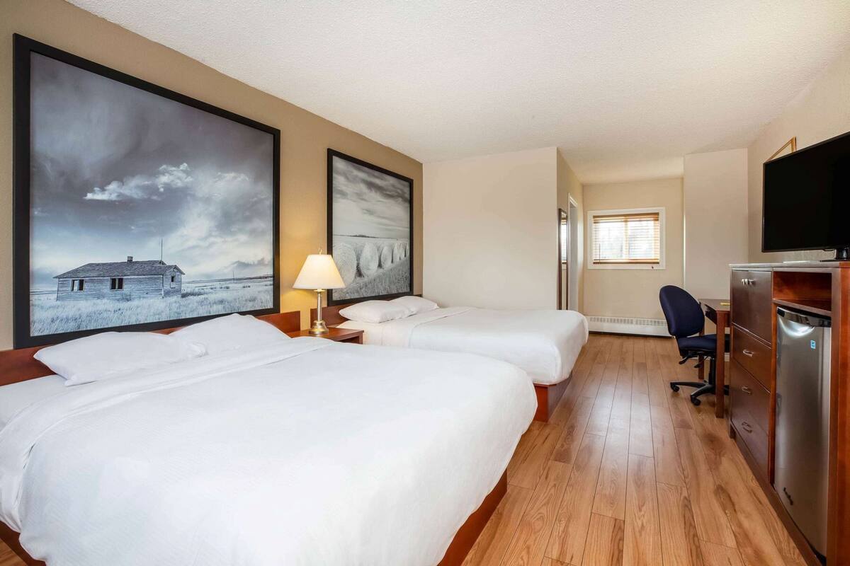 A hotel room with two beds, a desk, a chair, a TV, and artwork on the walls.