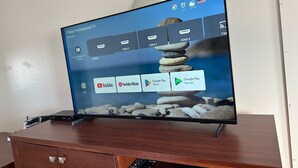 Televisor de pantalla plana sobre un soporte de madera con control remoto, mostrando una interfaz con iconos de aplicaciones y un widget del tiempo.