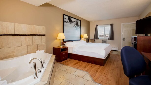 Habitación de hotel con una bañera grande, una cama con ropa blanca, dos lámparas de noche, un televisor de pantalla plana y una ventana con cortinas.