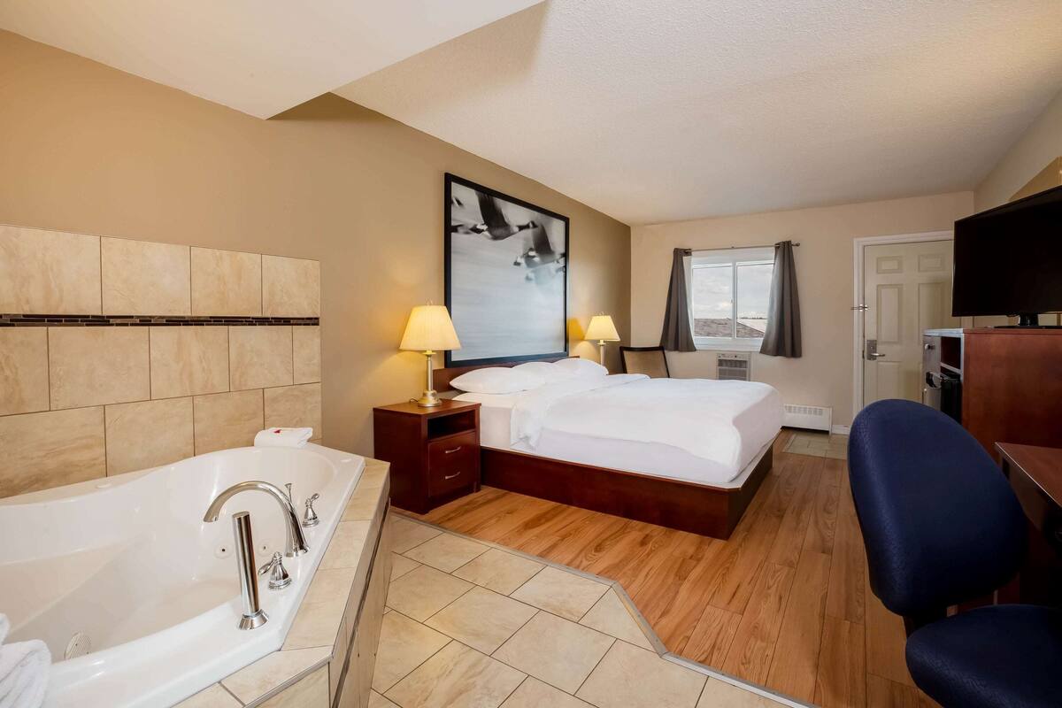 A hotel room with a large bathtub, a bed with white linens, two bedside lamps, a flat-screen TV, and a window with curtains.