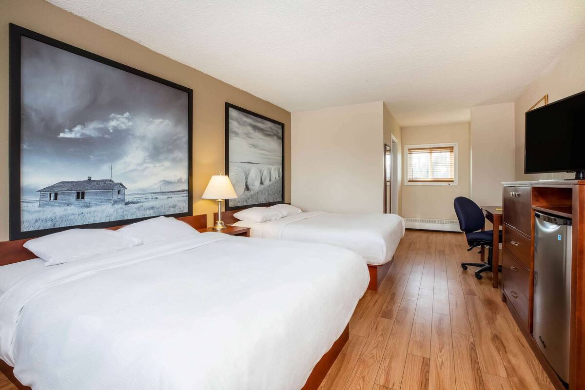 A hotel room with two beds, a desk, a chair, a TV, and artwork on the walls.