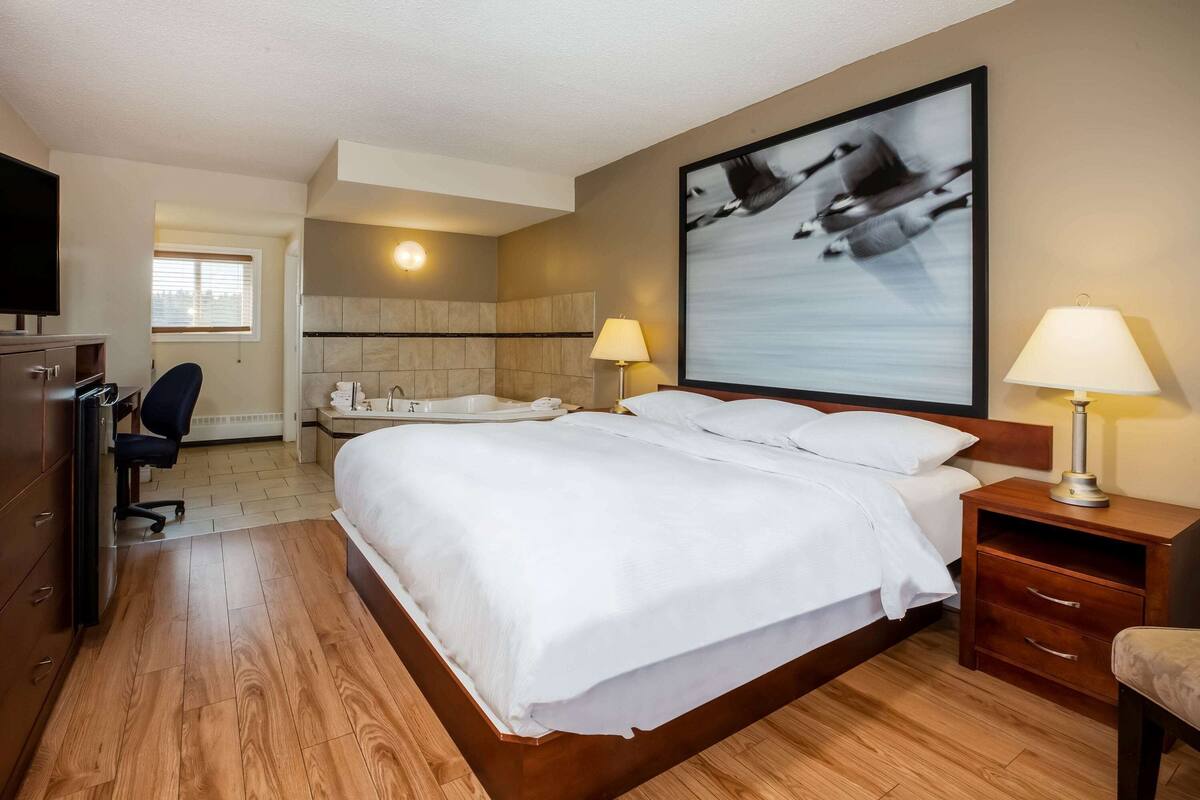 A hotel room with a large bed, a desk with a chair, a TV, a bathroom with a bathtub, and a wall-mounted artwork.