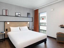 Habitación de hotel moderna con una cama grande, cabecera con iluminación integrada, una mesita de noche pequeña, una maleta y una ventana con vistas a un edificio.