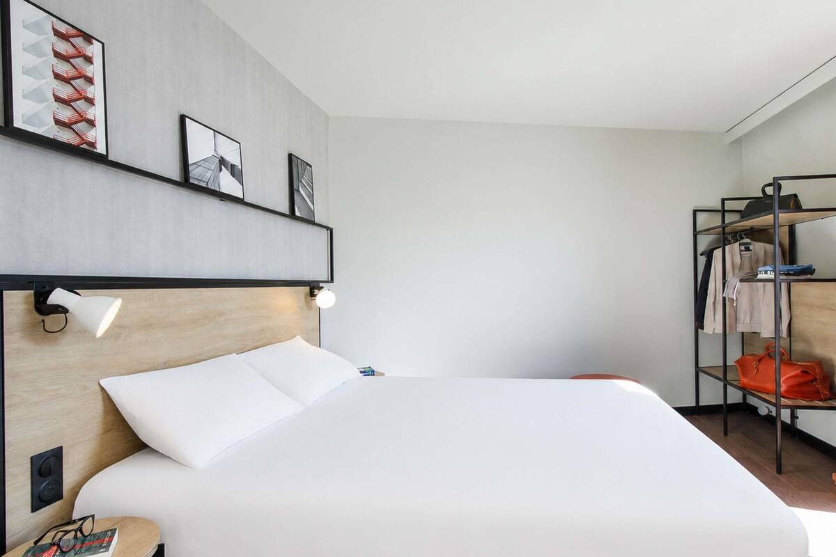 Quarto de hotel com uma cama grande, uma mesa de cabeceira com um livro, uma prateleira na parede com quadros e um cabideiro com malas.