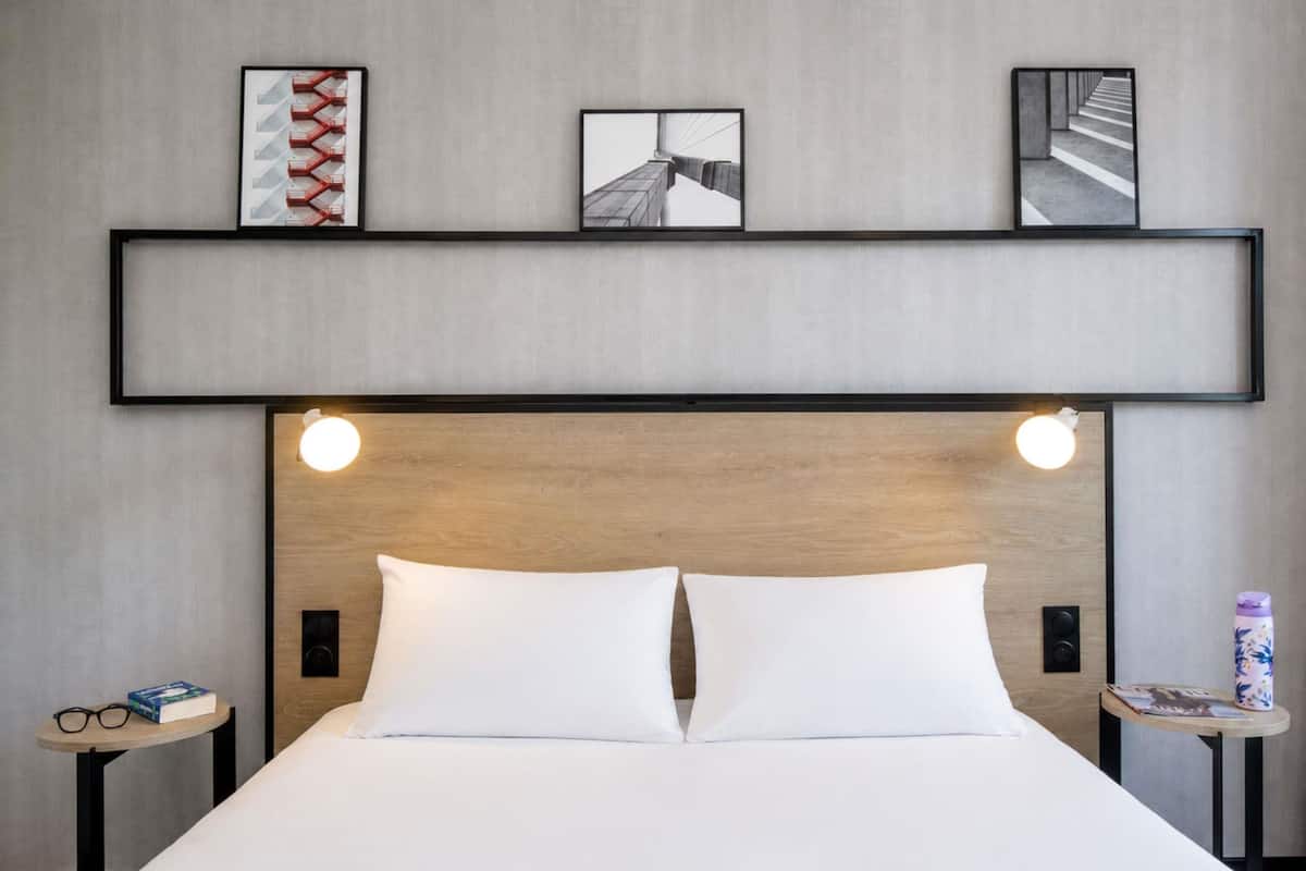 Quarto de hotel com cama, duas mesas de cabeceira, uma estante com quadros e cabeceira de madeira.