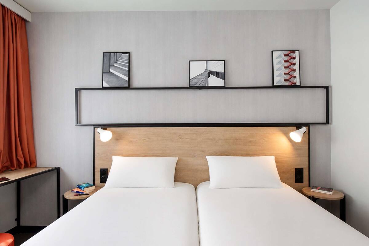 Quarto de hotel com cama de casal, duas mesas de cabeceira, cabeceira com iluminação embutida e três quadros na parede.