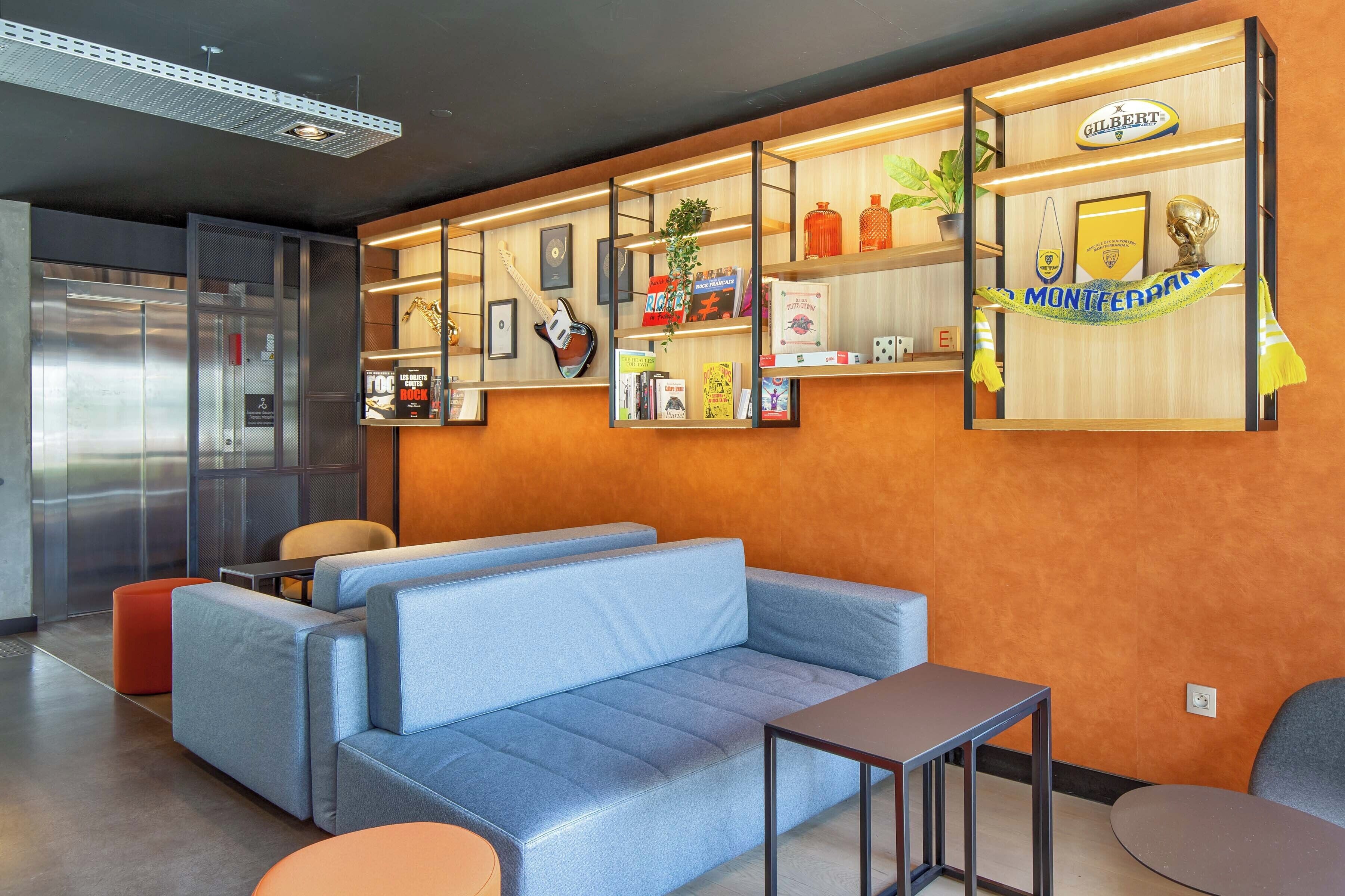 Un moderno vestíbulo de hotel con un sofá azul, una mesita pequeña y una estantería con varios objetos, incluyendo una planta, cuadros enmarcados y un letrero que dice 'MONTFERRAN'.