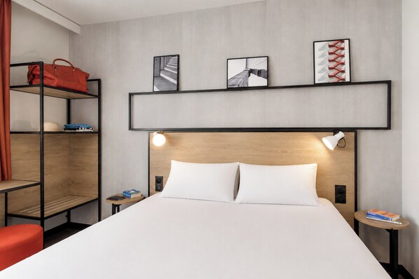 Una camera d'albergo con un letto, uno scaffale con una borsa rossa e tre quadri appesi al muro.