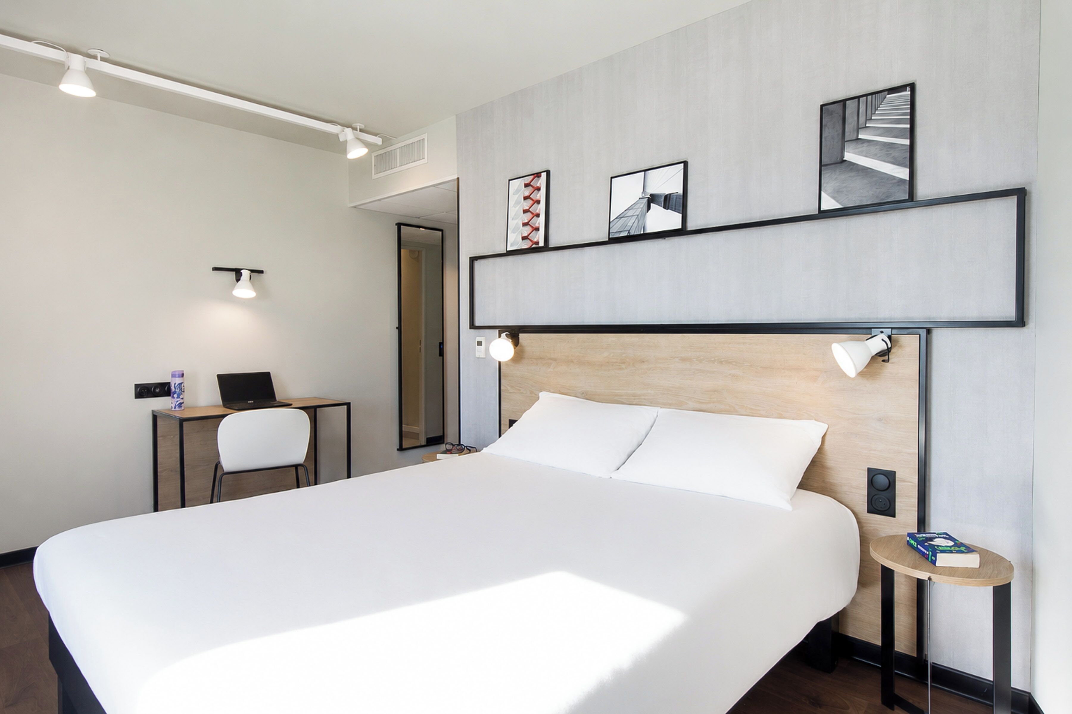 A modern hotel room with a large bed, a desk with a laptop, a chair, and a small table with a book.
