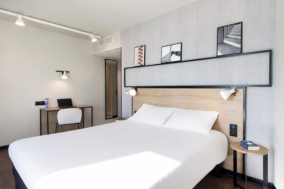 A modern hotel room with a large bed, a desk with a laptop, a chair, and a small table with a book.