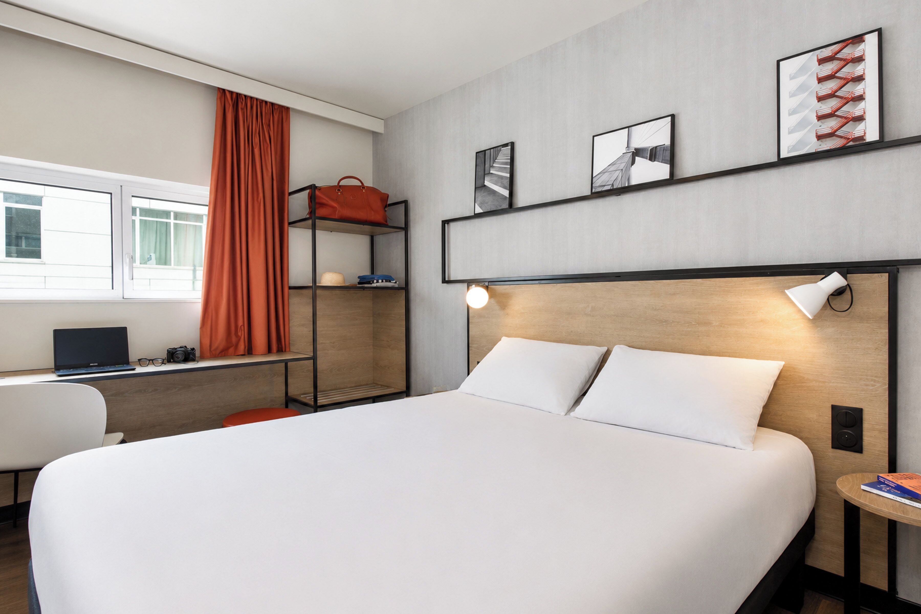 A modern hotel room with a large bed, a desk with a laptop, a chair, a shelf with a handbag, and artwork on the wall.