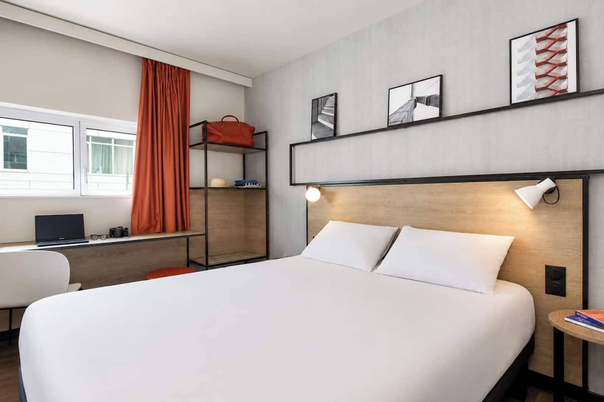 A modern hotel room with a large bed, a desk with a laptop, a chair, a shelf with a handbag, and artwork on the wall.