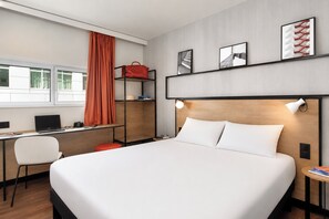 Una camera d'albergo con un letto grande, una scrivania con un portatile, una sedia, uno scaffale con una borsa rossa e un'opera d'arte al muro.