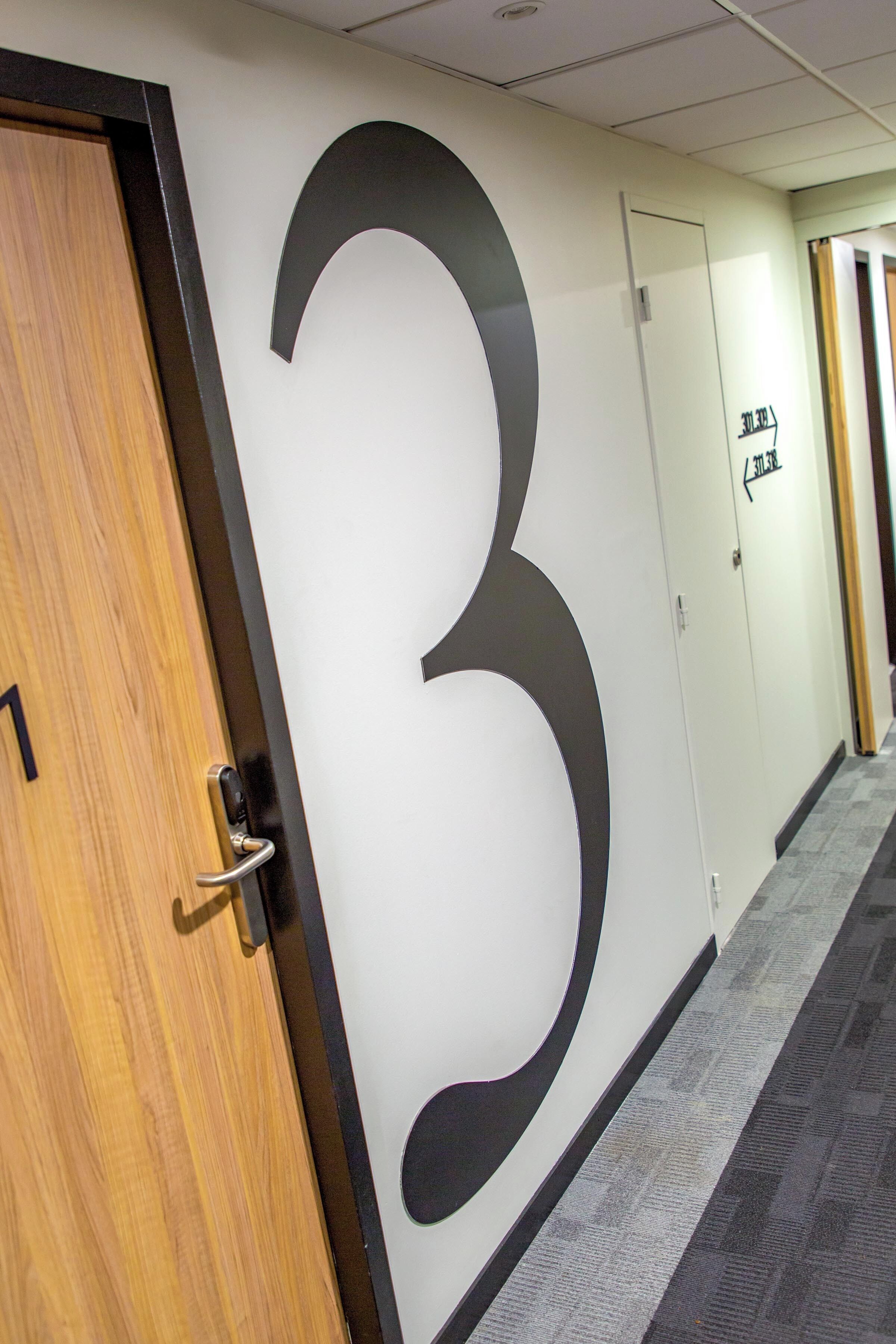 A hallway with a large number '3' on the wall, a wooden door with a metal handle, and a door with a sign above it.