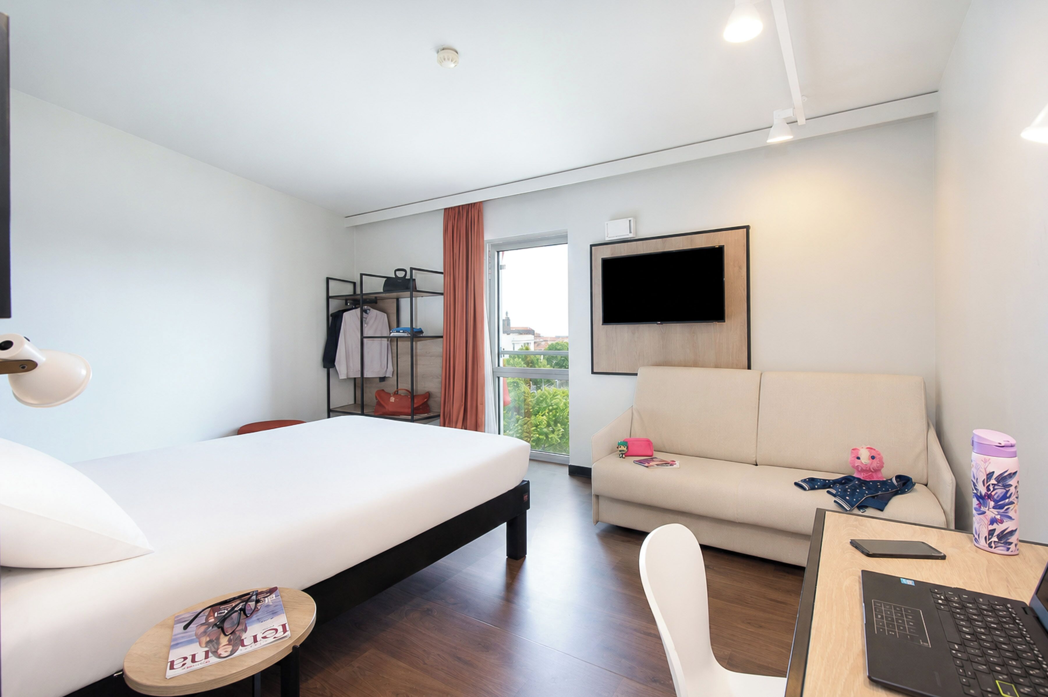 A modern hotel room with a bed, a sofa, a desk with a laptop, a TV mounted on the wall, and a window with a view.