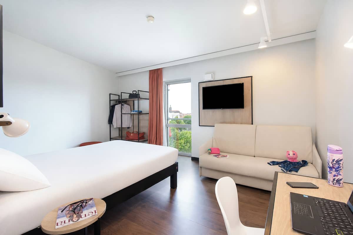 A modern hotel room with a bed, a sofa, a desk with a laptop, a TV mounted on the wall, and a window with a view.