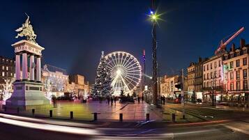 Una plaza urbana iluminada de noche, con una noria destacada, una estatua y edificios con luces.